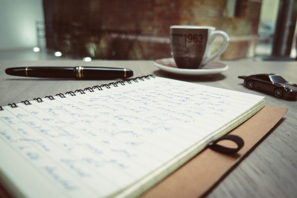 An open notebook with handwritten notes, a pen, and a cup of coffee on a desk.