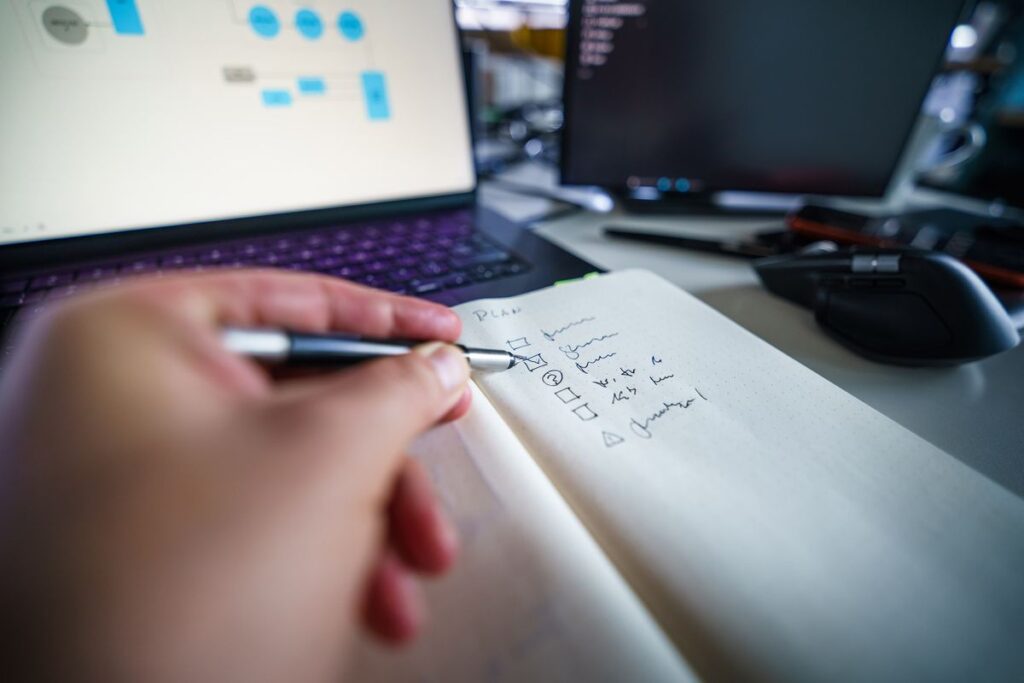 A person checks a handwritten checklist beside an open laptop at a desk.